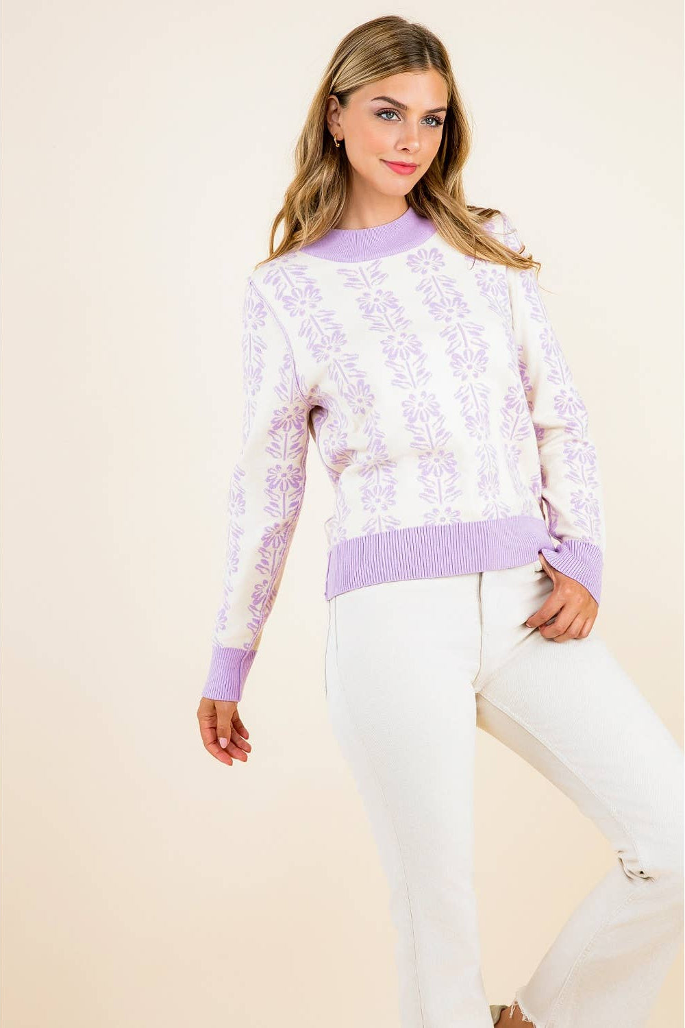 Woman wearing a light purple floral sweater and white pants on a beige background