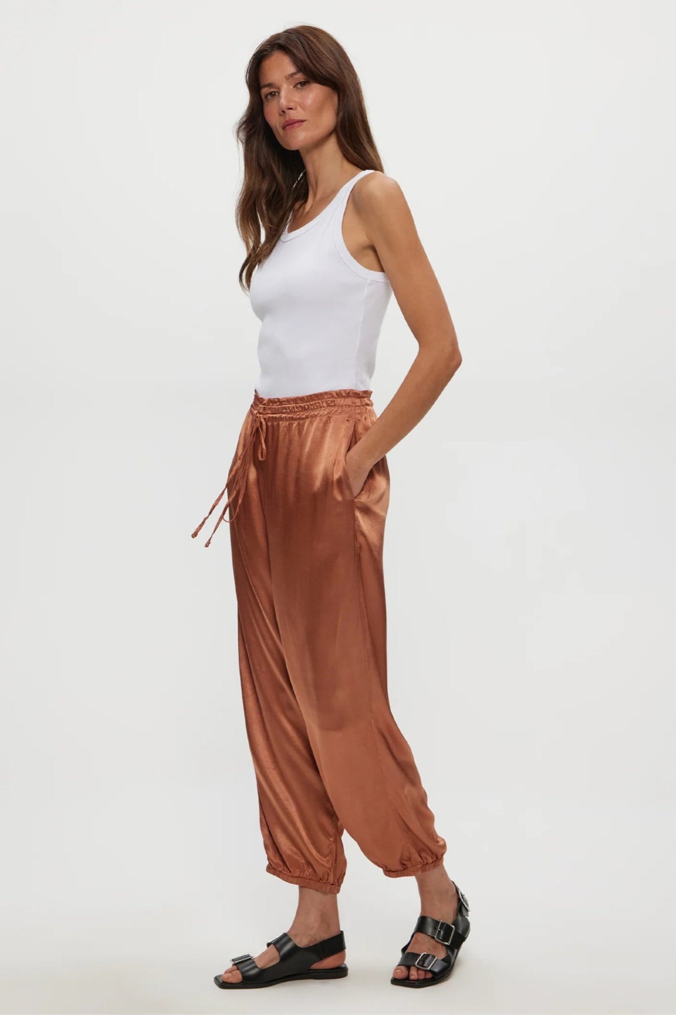 Woman wearing a white tank top and brown satin pants on a white background